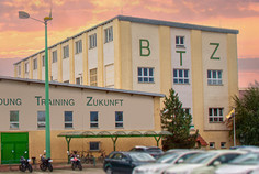 Das Bild zeigt das BTZ-Gebäude mit der Aufschrift „Bildung Training Zukunft“ bei Sonnenuntergang, im Vordergrund sind mehrere parkende Autos zu sehen.
