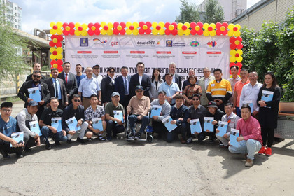 Gruppenfoto, darunter ein Mann im Rollstuhl, viele halten Zertifikate in der Hand, im Hintergrund eine Sponsorenwand mit mongolischer und deutscher Schrift sowie ein Blumenbogen aus Luftballons in Gelb und Rot.