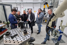 Ein Mann hält ein Schweißgerät in der Hand. Fünf Männer hören ihm aufmerksam zu , davor liegen verschiedene Werkzeuge und Bauteile auf einem Metalltisch.