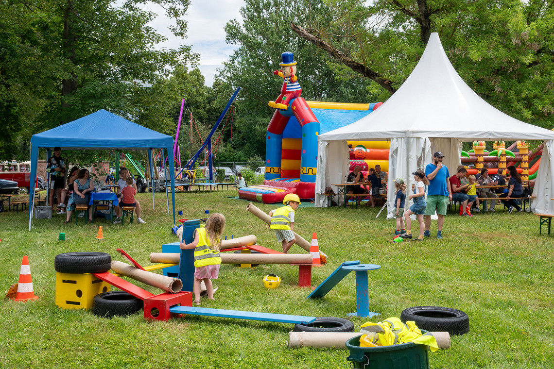 Im Vordergrund stehen und liegen bunte Spiel- und Bewegungsgeräte auf einer Wiese, wo zwei Kinder in gelber Warnweste spielen. Im Hintergrund befinden sich ein weißes Festzelt, ein blauer Pavillon, mehrere Personen sowie eine aufblasbare Spielfigur zwischen den Zelten.