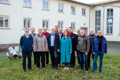 Zwölf Männer und zwei Frauen stehen in zwei Reihen auf einer Wiese vor einem hellen Institutsgebäude; alle blicken in die Kamera und tragen wetterfeste Kleidung.