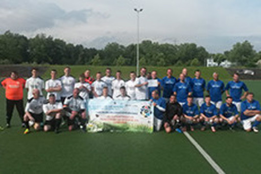 Eine Fußballmannschaft in weißen Trikots und eine in blauen Trikots posieren gemeinsam auf einem Sportplatz, in der Mitte halten sie ein Benefiz-Banner hoch.