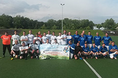 Eine Fußballmannschaft in weißen Trikots und eine in blauen Trikots posieren gemeinsam auf einem Sportplatz, in der Mitte halten sie ein Benefiz-Banner hoch.