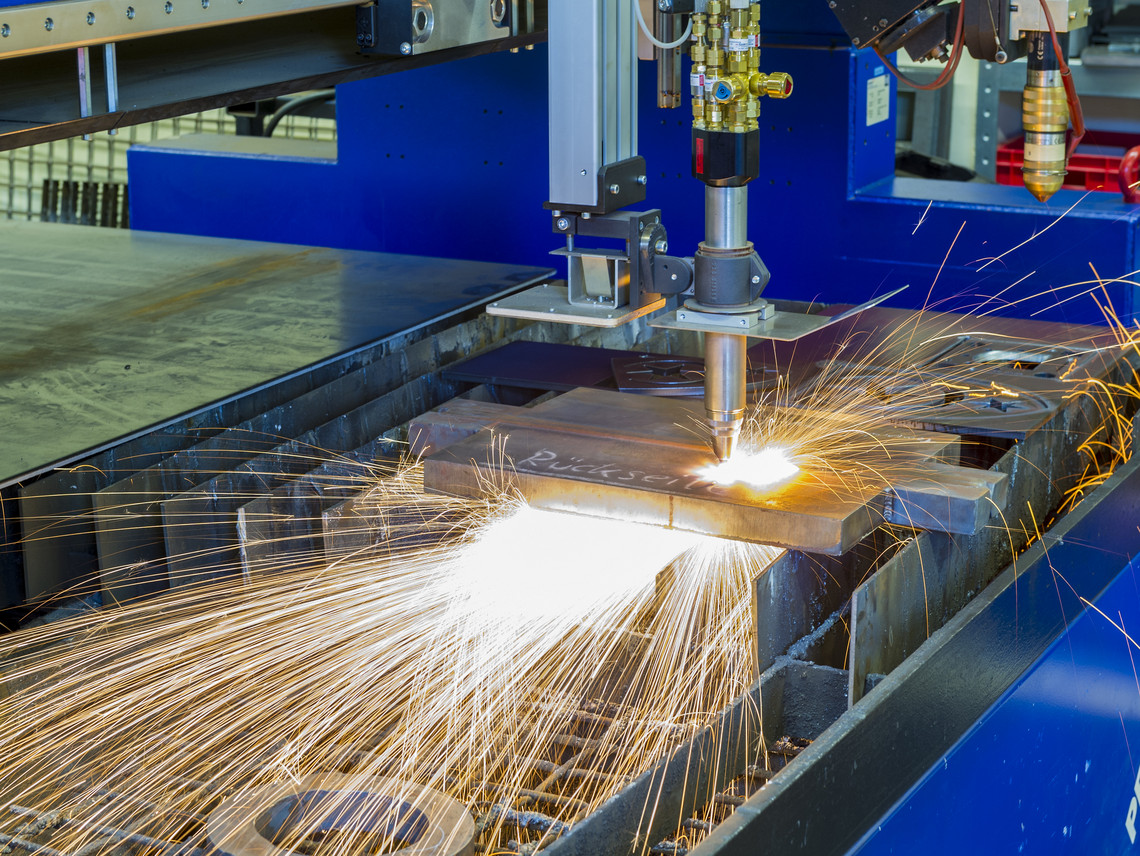 Automatisierte CNC-Schneidanlage schneidet mit einem Plasmastrahl durch eine dicke Metallplatte; helle Funken sprühen in weitem Bogen aus der Schnittstelle.