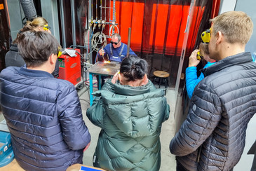 Vier Personen stehen in einer Werkstatt vor einem Schweißplatz und beobachten eine Schweißvorführung hinter roten Schutzvorhängen.