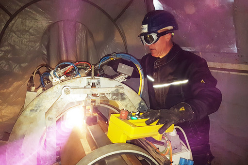 Eine Person in Schutzkleidung mit Helm und Schutzbrille bedient eine automatisierte Orbitalschweißanlage in einem Zelt, helles Licht tritt aus der Schweißstelle aus.