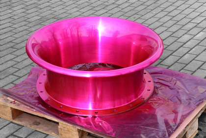 Eine glänzend pink eloxierte Einlauftrompete liegt auf einer Holzpalette mit schimmernder Folie, platziert auf einem gepflasterten Außenbereich.