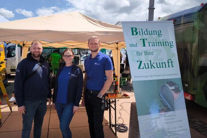 Zwei Männer und eine Frau stehen lächelnd vor einem Pavillon mit Messeequipment. Rechts im Bild steht ein aufgestelltes Banner mit dem Schriftzug „Bildung und Training für Ihre Zukunft“. Im Hintergrund sind Messfahrzeuge und Messinstrumente sichtbar.