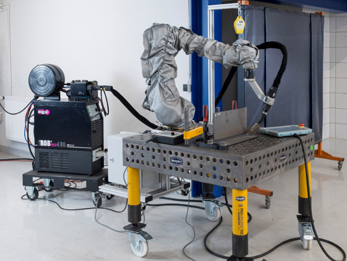Mobiler kollaborativer Roboterarm mit grauem Schutzüberzug führt einen Schweißprozess an einem Metallblech auf einem Lochrastertisch durch. Die Stromquelle steht auf einem Rollwagen.