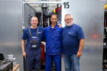 Drei Männer in blauer Arbeitskleidung stehen lächelnd in einer Werkhalle und posieren gemeinsam vor einer Schweißkabine mit der Nummer 15.