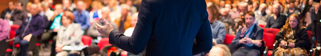 Ein Vortragender in dunklem Anzug spricht vor einem voll besetzten Auditorium mit rotem Gestühl; das Publikum hört aufmerksam zu, der Fokus liegt auf dem Rücken des Redners im Vordergrund.