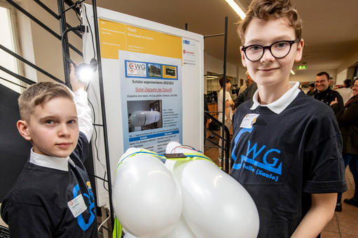 Zwei Jungen im schwarzen T-Shirt mit „WG Halle (Saale)“-Logo stehen vor einem Poster und halten ein Modell aus weißen Luftballons, im Hintergrund weitere Personen in einer Ausstellungshalle