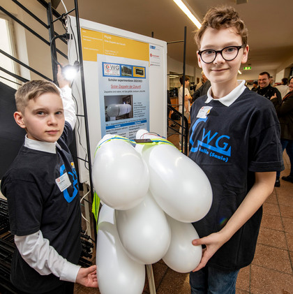 Zwei Jungen im schwarzen T-Shirt mit „WG Halle (Saale)“-Logo stehen vor einem Poster und halten ein Modell aus weißen Luftballons, im Hintergrund weitere Personen in einer Ausstellungshalle