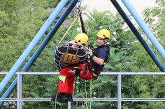 Zwei Personen mit Schutzhelmen hängen gesichert an Seilen und transportieren eine Person in einer Rettungstrage an einer Stahlkonstruktion, im Hintergrund sind Bäume sichtbar