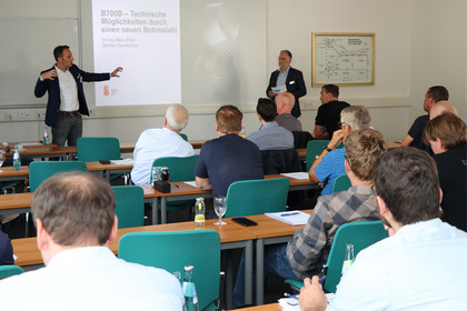 Mehr als zehn Personen sitzen an Tischen in einem Schulungsraum und blicken zu zwei Männern im vorderen Bereich. Einer der Männer steht an einem Rednerpult, der andere vor einer Tafel und zeigt auf die angezeigte Folie.