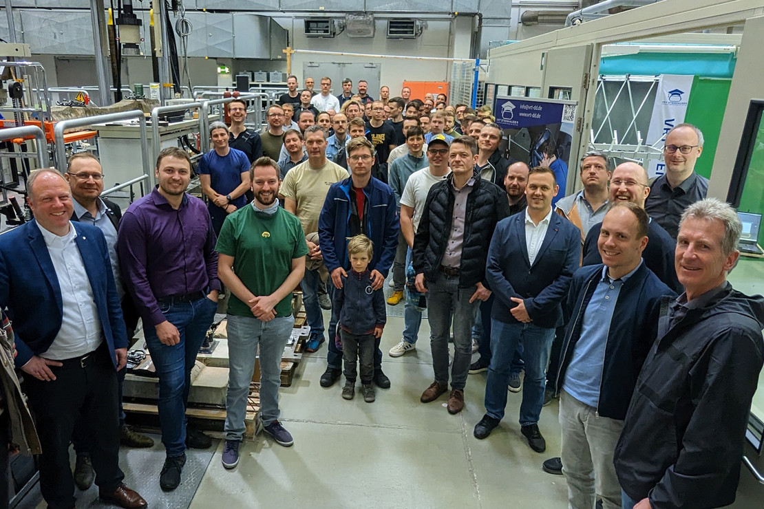 Gruppe von über 50 Personen steht lächelnd in einer Werkhalle, im Hintergrund Maschinen und technische Anlagen, vorn Männer im Business- und Freizeitlook
