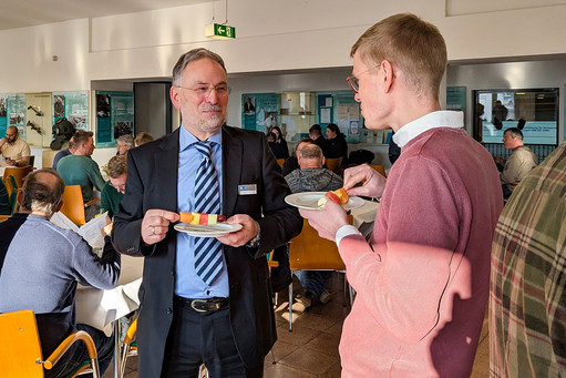 Mehrere Teilnehmende befinden sich im oberen Foyer des Veranstaltungszentrums, im Vordergrund unterhalten sich zwei Männer im Stehen und halten Teller mit Obst, im Hintergrund sitzen weitere Personen an Tischen und führen Gespräche oder arbeiten mit Unterlagen