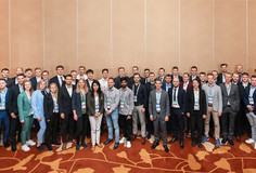 Gruppenfoto von jungen Erwachsenen in Businesskleidung mit Namensschildern, aufgenommen in einem Konferenzraum mit gemustertem Teppich und beiger Wandverkleidung.