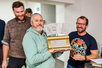 Drei Männer stehen lächelnd zusammen, einer hält eine Holzbox mit Pokalen in den Händen, im Hintergrund weißer Raum.