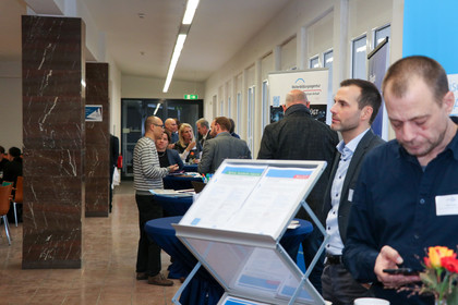 Teilnehmende einer Fachveranstaltung stehen an Infoständen in einem Flur mit Marmorsäulen und großen Fenstern; im Vordergrund Informationsmaterial auf einem Aufsteller und Personen im Gespräch.