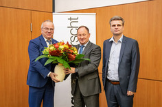 Drei Männer in Anzügen stehen vor einer Holzvertäfelung und einem Banner mit dem Wort „Wirtschaft“, zwei von ihnen halten gemeinsam ein herbstlich arrangiertes Blumenbouquet in einer Vase.