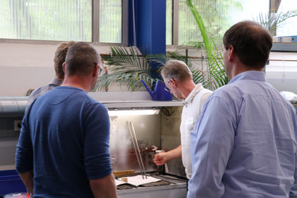 Drei Männer stehen an einem Prüfaufbau mit einem metallischen Bauteil und betrachten den Arbeitsbereich. Einer der Männer arbeitet mit einem Werkzeug am Bauteil, im Hintergrund sind technische Geräte und Fenster zu sehen.