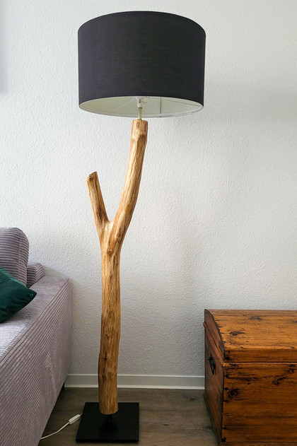 Eine Stehlampe aus einer naturbelassenden 1,5 m hohen Astgabel in einem Wohnzimmer. Links daneben ist ein graues Sofa mit einem grünen Kissen, rechts eine rustikale Holzkiste.