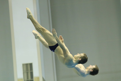 Zwei Wasserspringer führen synchron einen Sprung aus und befinden sich in gespannter Körperhaltung kopfüber in der Luft; der Hintergrund zeigt die Innenwand der Schwimmhalle.