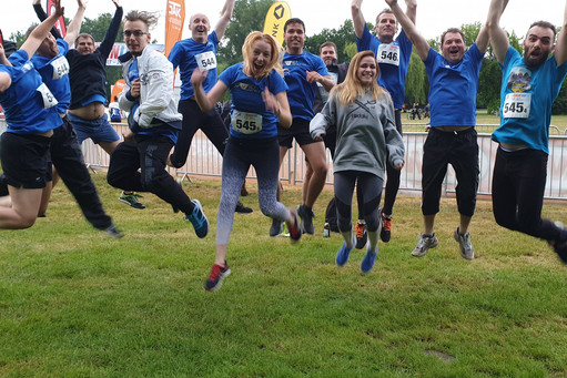 Neun Männer und drei Frauen springen mit Startnummern lachend in die Luft, alle tragen Sportkleidung. Im Hintergrund sind Absperrgitter, Banner und weitere Teilnehmende eines Laufs zu sehen.