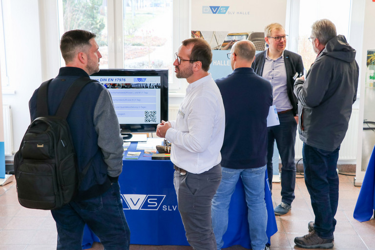 Mehrere Personen stehen an einem Informationsstand der SLV Halle GmbH mit Monitor und tauschen sich über Inhalte aus