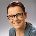 Katarina Schmit, in grauem Anzug mit Brille, lächelnd vor neutralem Hintergrund