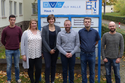Sechs Personen, vier Männer und zwei Frauen, stehen nebeneinander vor einem Wegweiser der SLV Halle mit DVS-Logo. Sie posieren für ein Gruppenfoto, im Hintergrund sind ein Gebäude und herbstlich gefärbte Bäume zu sehen.