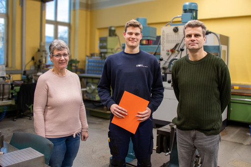 Junger Mann in Arbeitskleidung hält orangefarbene Mappe, steht zwischen einer Frau im rosa Pullover und einem Mann im grünen Pullover in einer Werkstatt mit Maschinen im Hintergrund