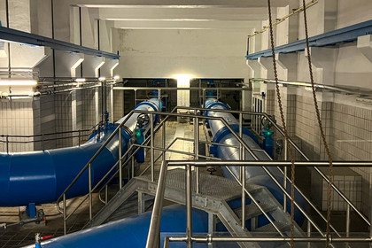 Technikraum mit mehreren großen blauen Rohrleitungen, Metallgeländern und gefliesten Wänden; die Anlage ist hell ausgeleuchtet und vermittelt den Eindruck eines Wassersystems.