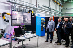 Fünf Männer betrachten auf einem großen Monitor die Aufnahme eines technischen Bearbeitungsprozesses in einer Werkhalle.
