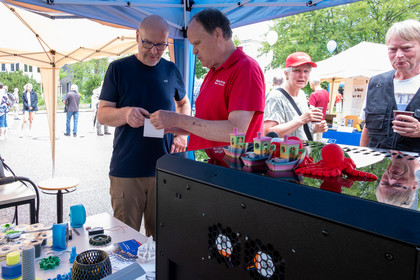 Vier Personen stehen an einem Stand unter einem Pavillon und unterhalten sich einen 3D-Drucker. Auf dem Tisch stehen Blumen in Vasen und weitere Dekorationsobjekte aus dem 3D-Drucker, im Hintergrund sind weitere Marktstände und Besucher zu sehen.