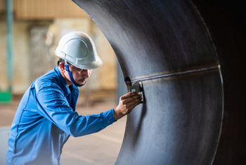 Arbeiter mit Schutzhelm und blauer Kleidung inspiziert die Innenseite eines großen Metallrohrs mit einem Prüfgerät.