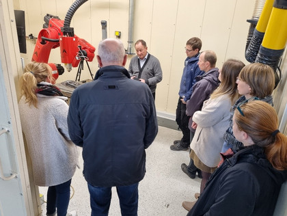 Acht Personen, darunter fünf Frauen und drei Männer, stehen in einem Technikraum um einen Mann, der eine rote Schweißroboteranlage erklärt; alle blicken konzentriert auf das Bauteil in seinen Händen.