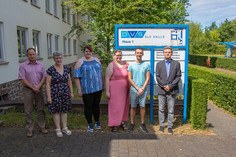 Sechs Personen stehen bei sonnigem Wetter nebeneinander vor einem blauen Wegweiser der DVS SLV Halle auf dem Gelände zwischen Gebäude und Hecke und blicken in die Kamera.