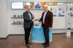 Zwei Männer im Anzug stehen im Veranstaltungsfoyer der SLV Halle GmbH und übergeben sich symbolisch einen blauen Staffelstab. Im Hintergrund sind Broschüren und eine Infotafel mit Leistungen wie Aus- und Weiterbildung, Qualitätssicherung und Forschung sichtbar.