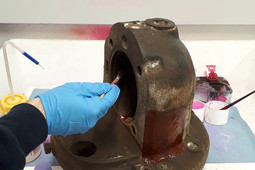 Eine Hand mit blauer Schutzhandschuhen trägt mit einem Pinsel rotes Farbeindringmittel auf ein großes Metallbauteil auf; im Hintergrund stehen Behälter mit weiteren Prüfmitteln.