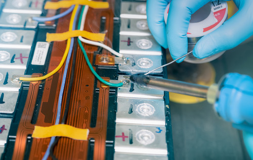 Nahaufnahme einer Person mit blauen Schutzhandschuhen beim Löten an einem Batteriepack, mehrere Zellen, Kabel und flexible Leiterbahnen sind sichtbar.