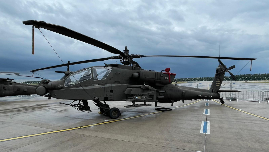Militärhubschrauber vom Typ Apache steht auf nassem Rollfeld, Rotorblätter quer ausgerichtet, Bewaffnung sichtbar, dunkle Wolken im Hintergrund