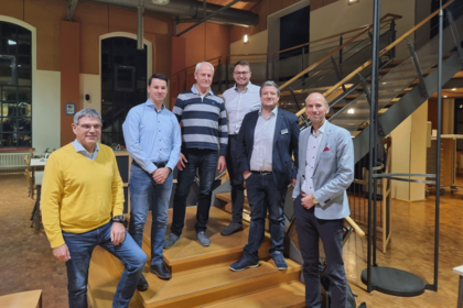 Sechs Männer stehen locker gruppiert auf einer Holztreppe in einem modern beleuchteten Raum mit industriellem Charme und Metalltreppe im Hintergrund, sie blicken lächelnd in die Kamera.