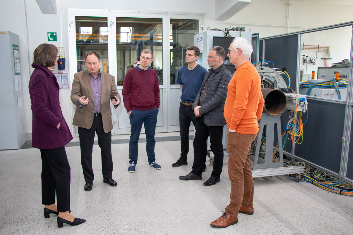 Sechs Personen stehen in einer Werkhalle der SLV Halle GmbH in einer Gesprächsrunde. Eine Person spricht gestikulierend, während die anderen aufmerksam zuhören. Im Hintergrund sind technische Geräte und Kabel zu sehen.