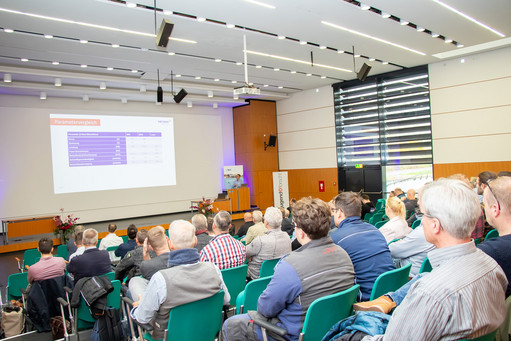 Publikum sitzt in einem Konferenzsaal und hört einem Vortrag zu, eine Präsentation zeigt Schweißparameter im Vergleich