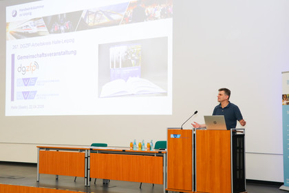 Eine Person steht an einem Rednerpult in einem Hörsaal und spricht vor einer projizierten Präsentationsfolie. Im Vordergrund stehen mehrere Tische mit Geräten, im Hintergrund ist eine große Leinwand mit einer Folie zu sehen.