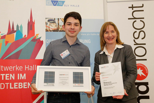 Ein junger Gewinner präsentiert stolz seinen Preis beim Wettbewerb "Jugend forscht". Neben ihm steht eine Frau mit einer Urkunde, beide lächeln in die Kamera. Im Hintergrund sind Logos der Stadt Halle, Fraunhofer und SLV Halle zu sehen.