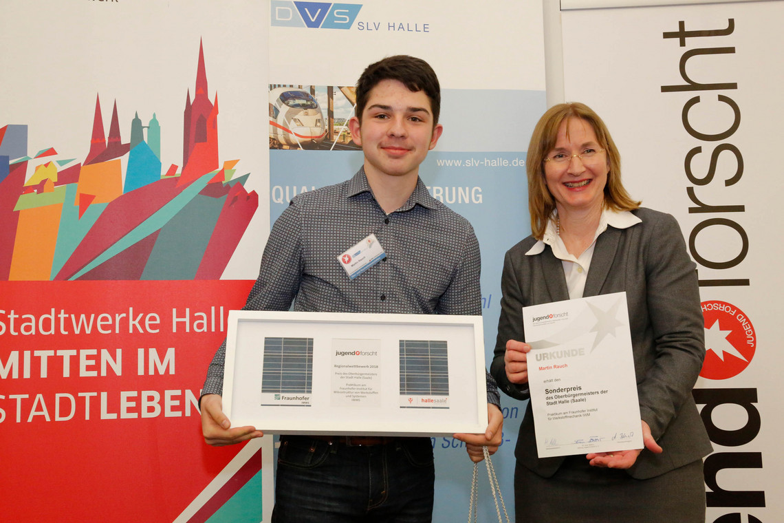 Ein junger Gewinner präsentiert stolz seinen Preis beim Wettbewerb "Jugend forscht". Neben ihm steht eine Frau mit einer Urkunde, beide lächeln in die Kamera. Im Hintergrund sind Logos der Stadt Halle, Fraunhofer und SLV Halle zu sehen.