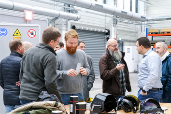 Gruppe von Männern in einer Werkhalle im Gespräch, einige halten Metallteile, auf dem Tisch liegen Schutzausrüstung wie Helme und Schutzbrillen, Warnschilder an der Wand.
